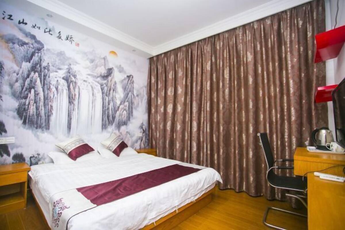 A hotel room with a bed, curtains, a desk with a chair, and a wall with a painting.