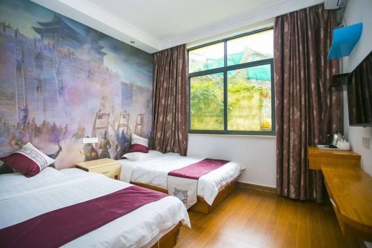 A hotel room with two beds, a large window with curtains, a mural on the wall, and a wooden floor.
