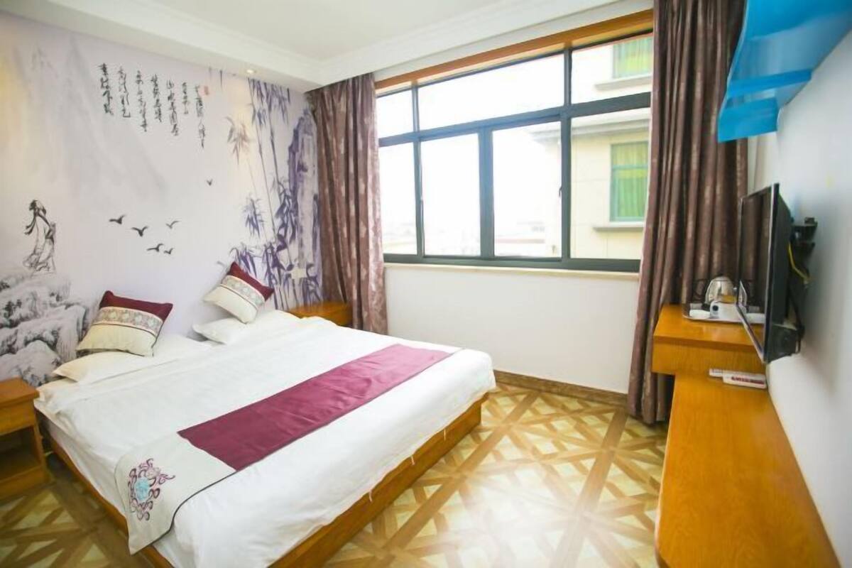 A hotel room with a bed, a TV, a window, and a wall with artwork.