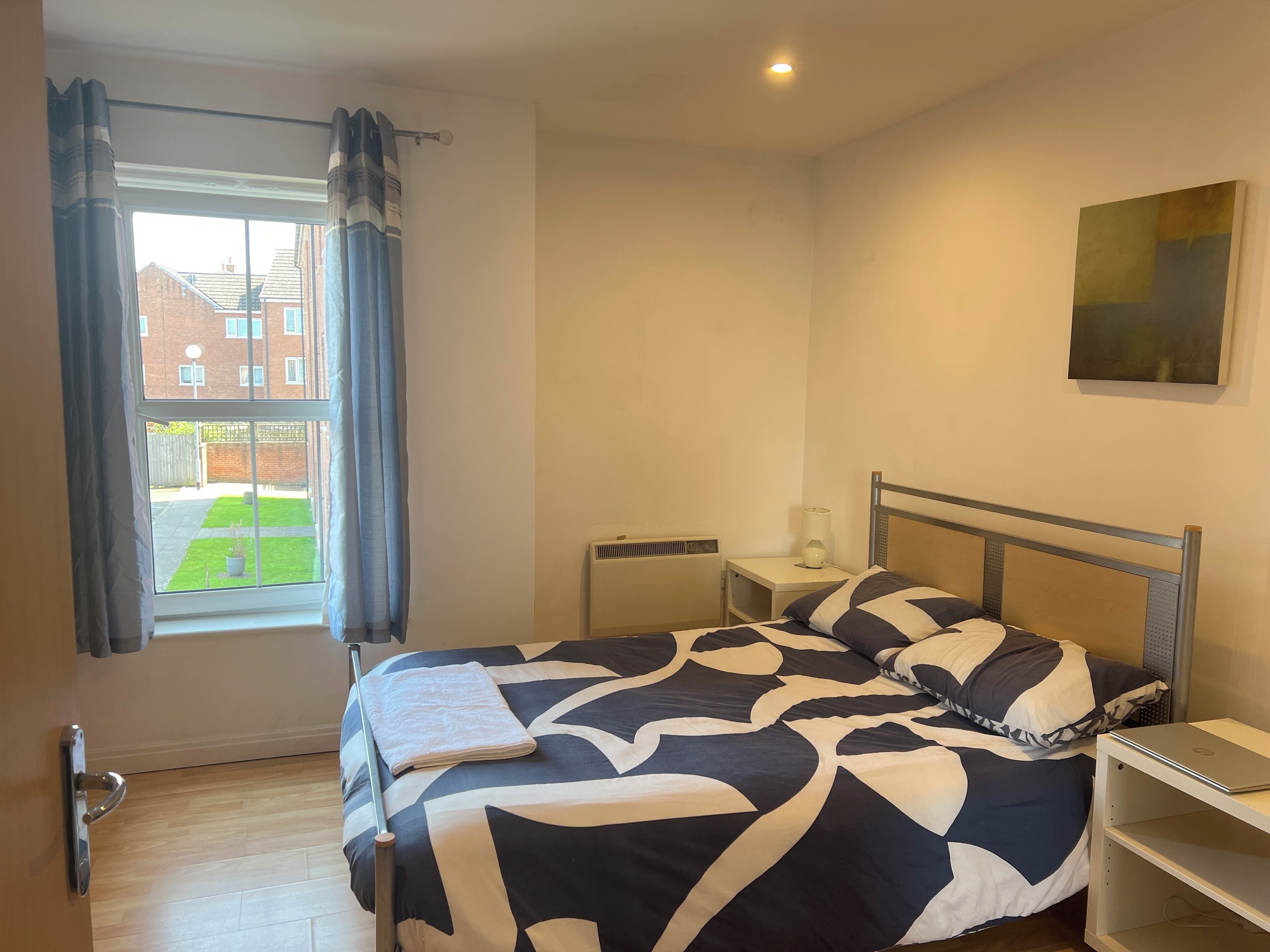A bedroom with a bed, a nightstand, a radiator, a window with curtains, and a view of a building outside.