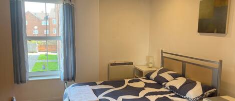 A bedroom with a bed, a nightstand, a radiator, a window with curtains, and a view of a building outside.