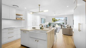 A modern kitchen with white cabinetry, a central island with a sink, and a dining area with a sofa and chairs. - Key West Beach House (Coolum Beach)