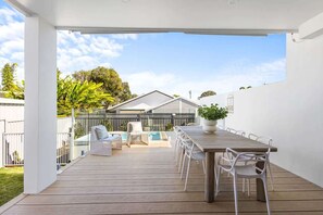 An outdoor patio with a wooden deck, white chairs, a dining table, and a pool. - Key West Beach House (Coolum Beach)