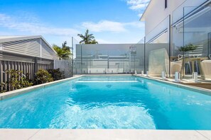A rectangular swimming pool with clear blue water, surrounded by a white fence and a modern building with large windows. - Key West Beach House (Coolum Beach)