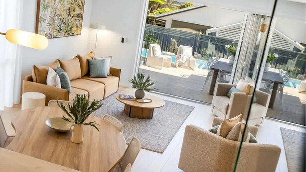 A modern living room with a sofa, coffee table, and dining area, a glass door leading to a poolside area. - Key West Beach House (Coolum Beach)