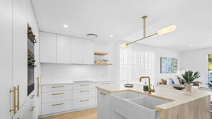 A modern kitchen with white cabinetry, gold handles, a central island with a sink, and pendant lighting. - Key West Beach House (Coolum Beach)
