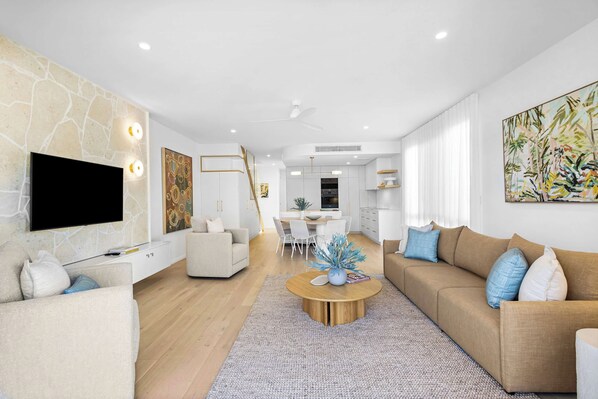 A modern living room with a flat-screen TV, a sofa, a coffee table, and a dining area with a table and chairs.