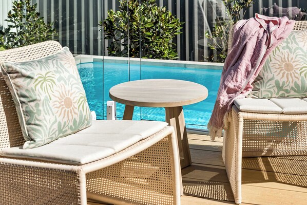 A poolside seating area with wicker chairs and cushions, a small round table, and a view of a swimming pool.