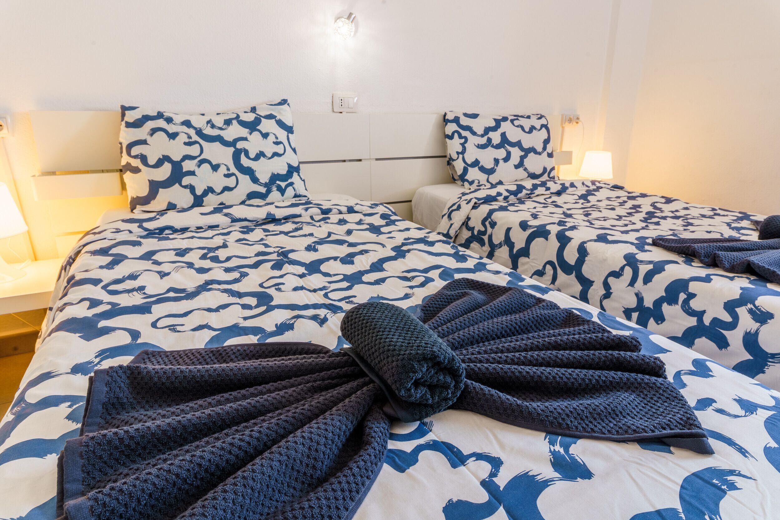 Quarto de hotel com duas camas, cada uma com colcha e almofadas estampadas em azul e branco.