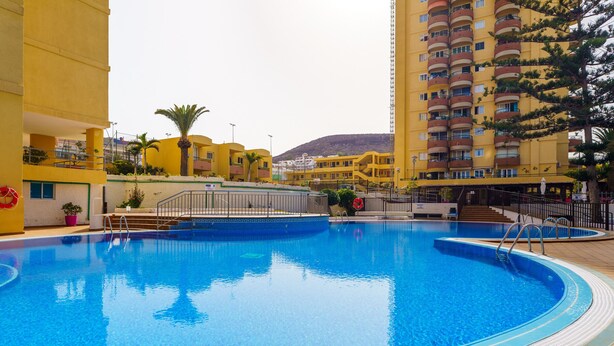 A swimming pool surrounded by yellow buildings with balconies, a palm tree, and a mountain in the background. - Suntower Arona - Seaviews & Pool - next to beach (Arona)