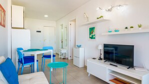 A modern living room with a TV, white furniture, and a dining area with blue chairs. - Suntower Arona - Seaviews & Pool - next to beach (Arona)