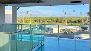 A spacious balcony with a glass railing overlooking a construction site and palm trees. - CoHost Flat Porto Plaza 409 (IPOJUCA)