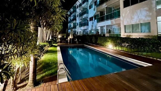 A swimming pool with a wooden deck, surrounded by greenery and a modern building with balconies. - CoHost Flat Porto Plaza 409 (IPOJUCA)