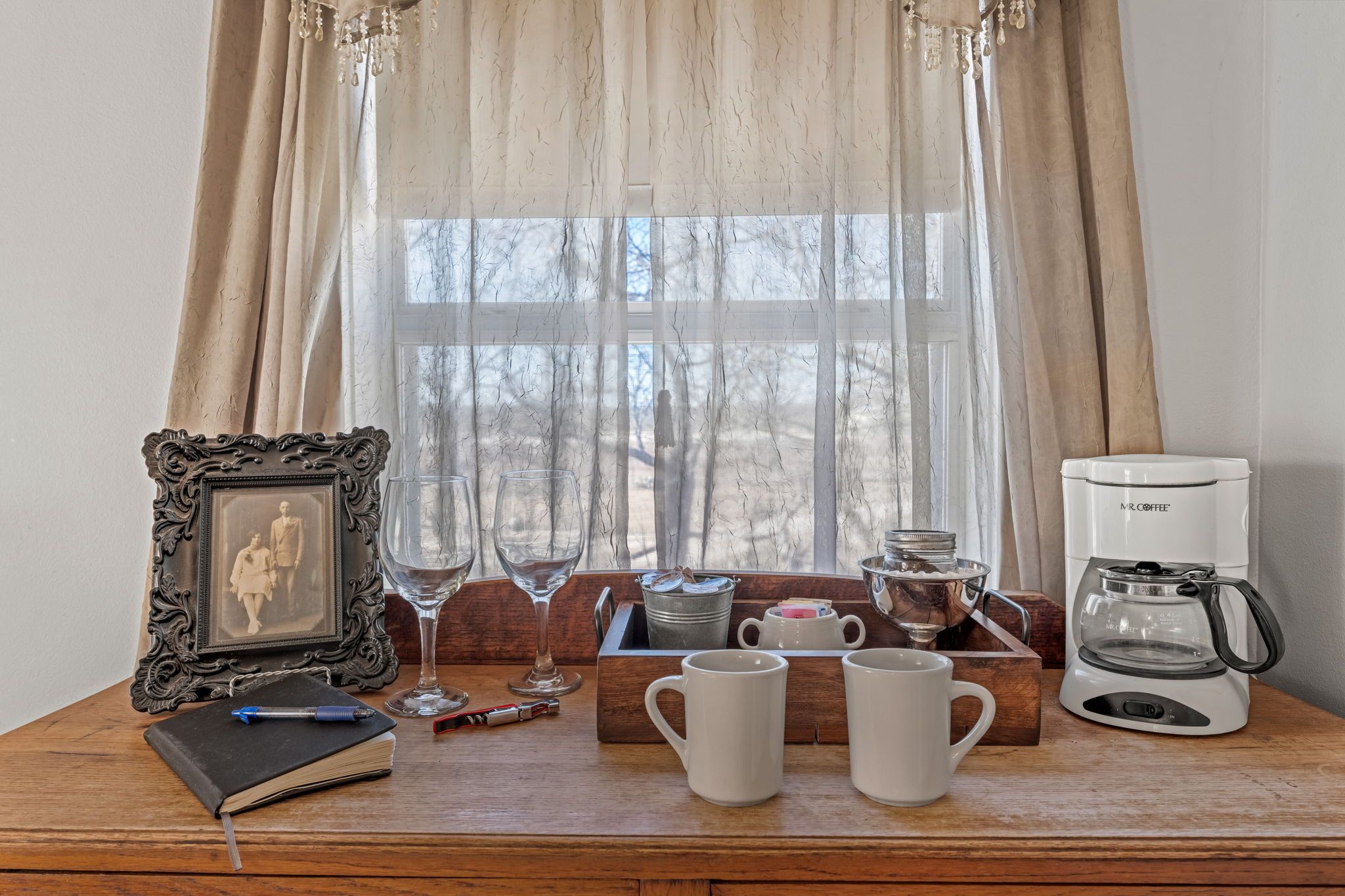 A wooden table with a framed picture, a notebook, a pen, two wine glasses, a mug, a coffee maker, and a kettle.