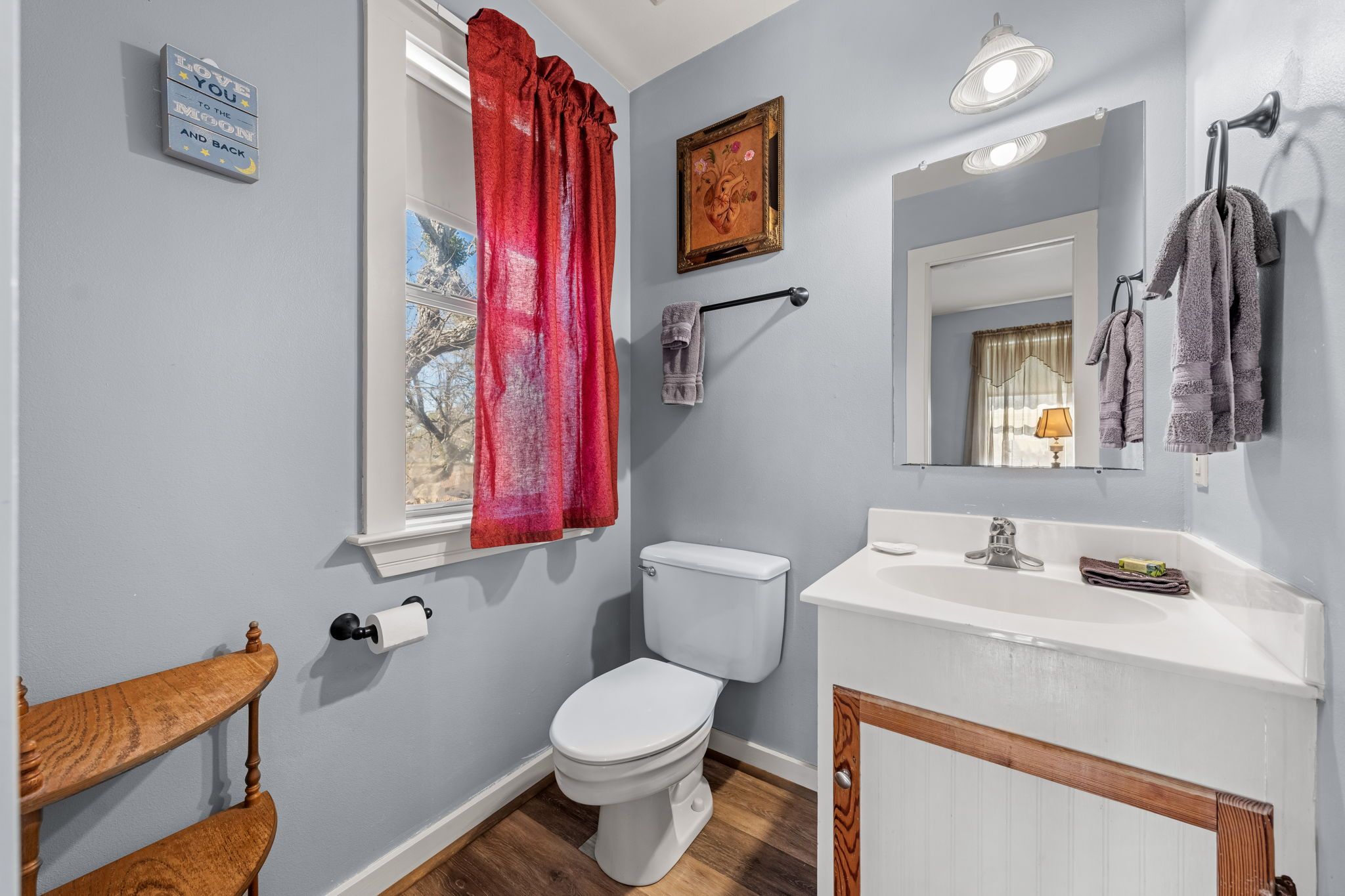 A bathroom with a toilet, sink, mirror, towel rack, and a window with a red curtain.