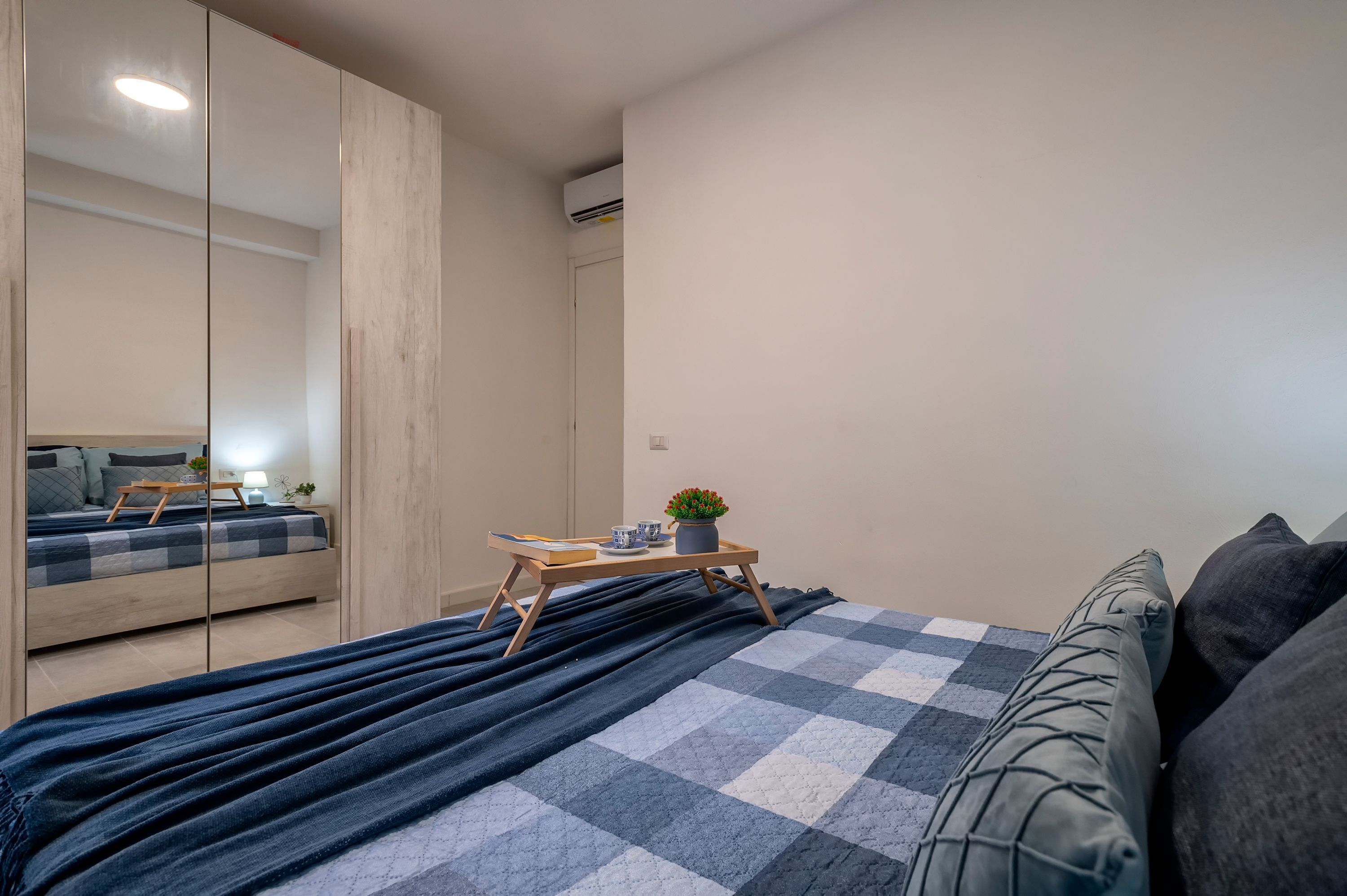 A bedroom with a bed, a bedside table with a lamp and a plant, a mirror, and a wardrobe.