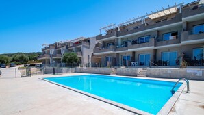 A swimming pool with a clear blue water, surrounded by a concrete deck, and adjacent to a multi-story building with balconies and windows. - Sardinia Re - Armonia 9 (Castelsardo)