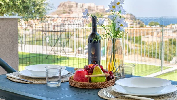 A table set with plates, a bottle of wine, a glass of water, a bowl of fruit, and a vase with flowers, overlooking a scenic view. - Sardinia Re - Armonia 9 (Castelsardo)