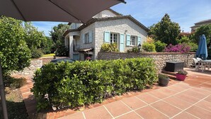 Ein Steinhhaus mit einer gefliesten Terrasse, ein Auto davor und ein Hecke im Vordergrund.