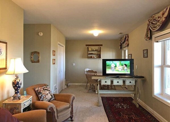 Una sala de estar con televisor, una silla, una mesita con lámpara y un ventanal.