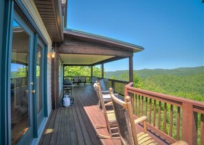 Una terraza de madera con mobiliario exterior, una parrilla y una vista panorámica de una cadena montañosa boscosa.
