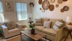 Un salon avec un canapé beige, une table basse en bois et un coussin décoratif à motifs. On y trouve une lampe fixée au mur et une petite plante en pot posée sur la table basse.
