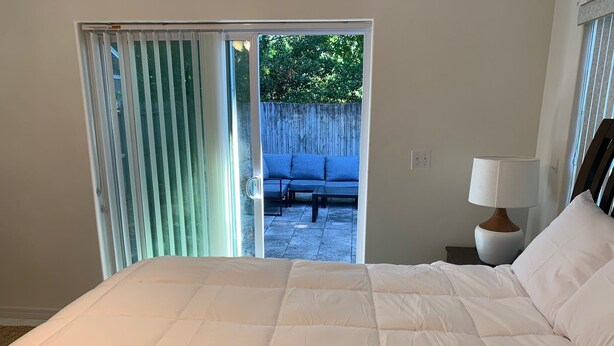 Une chambre dotée d’un grand lit, d’une table de chevet avec une lampe et offrant une vue sur un espace extérieur aménagé, accessible par une porte coulissante.