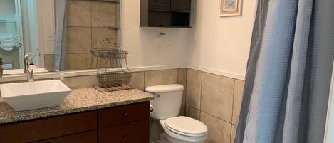 Un baño con lavamanos blanco, mueble de tocador de madera oscura, inodoro y cortina de ducha.