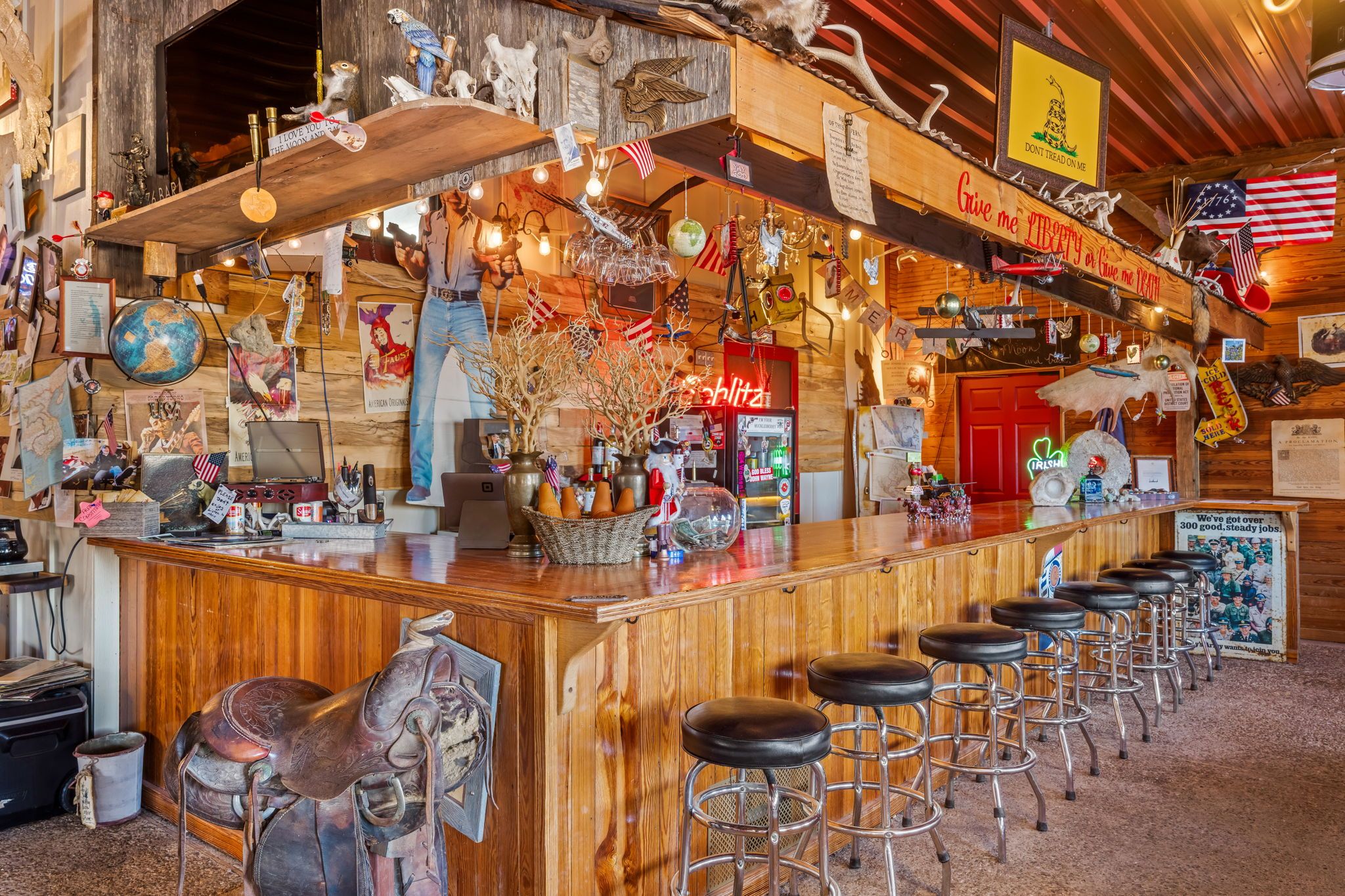 Uma área de bar com balcão de madeira, banquetas e várias decorações, incluindo bandeiras e um letreiro de neon.