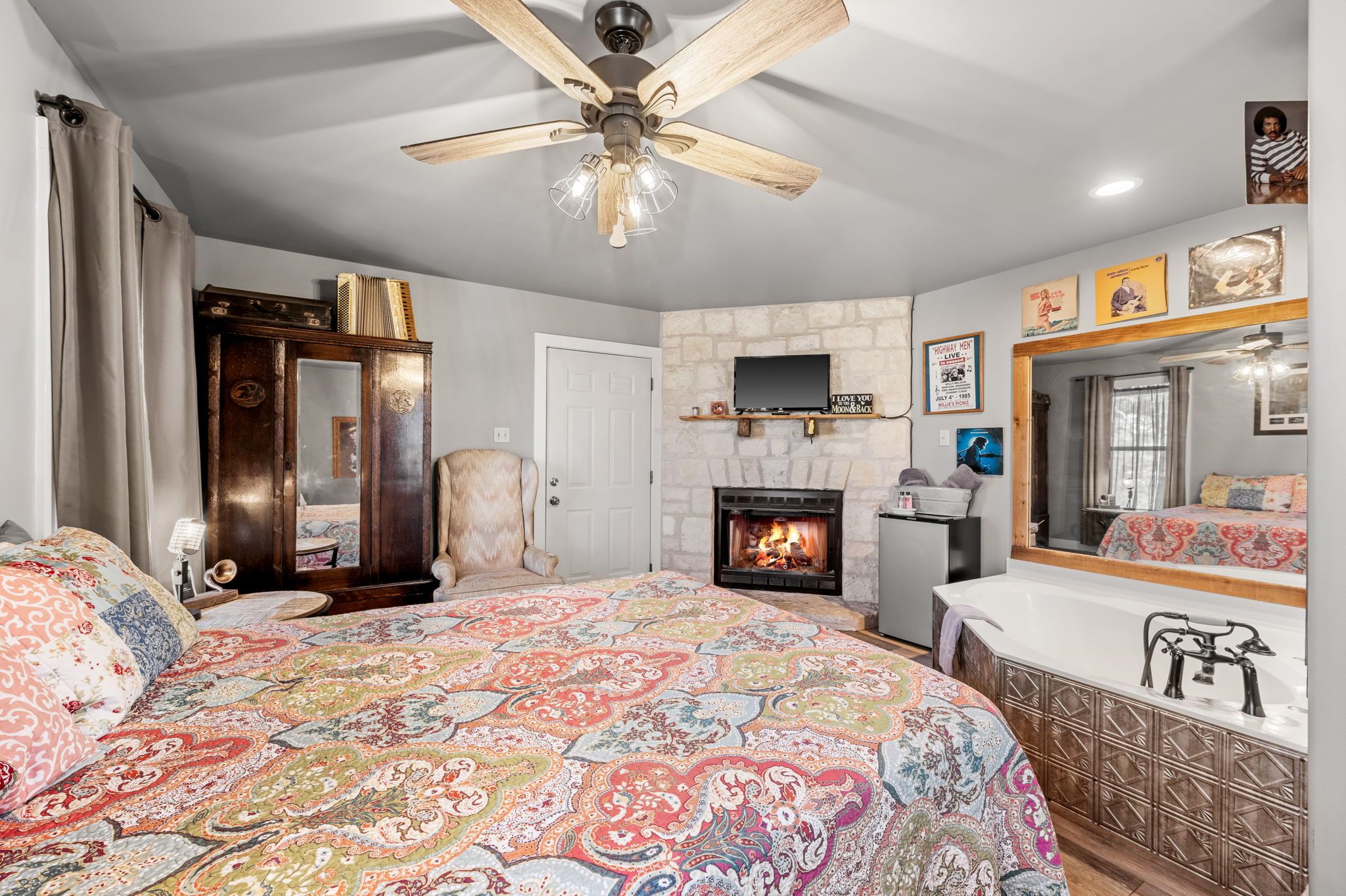 A bedroom with a large bed, a fireplace, a ceiling fan, a TV, and a bathroom with a bathtub.