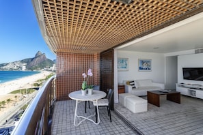 A balcony with a view of the beach and mountains, a white sofa, a small table with chairs, and a TV. - 801 (Rio de Janeiro)