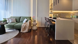 Ein modernes Wohnzimmer mit einem grünen Sofa, einem runden Couchtisch und einem kleinen Beistelltisch mit einer Vase. Ein Küchenbereich mit einer weißen Kochinsel und Barhocker ist ebenfalls zu sehen.