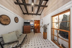 Un intérieur chaleureux avec un canapé, des poutres en bois au plafond, une enseigne murale représentant du vin et de la bière, un escalier et une fenêtre où sont exposés des vêtements.