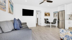 Una sala moderna con un sofá gris, un comedor con mesa y sillas blancas, una cocina con gabinetes blancos y un refrigerador de acero inoxidable, y un ventilador de techo.