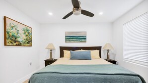 Un dormitorio con cama, dos mesitas de noche con lámparas, ventilador de techo, un cuadro en la pared y una ventana con persianas.