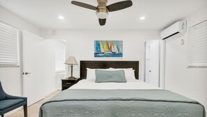 A bedroom with a bed, a ceiling fan, a nightstand, a lamp, and a painting of sailboats.