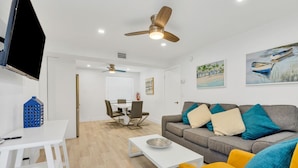 Una sala moderna con un sofá gris, un sillón amarillo, una mesa de centro blanca y un televisor montado en la pared. Hay una zona de comedor con mesa y sillas, y un ventilador de techo.