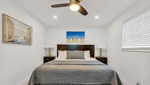 A bedroom with a bed, two bedside tables, a ceiling fan, a painting, and a window with blinds. - Cozy two bedroom, feet from the beach (Hollywood)