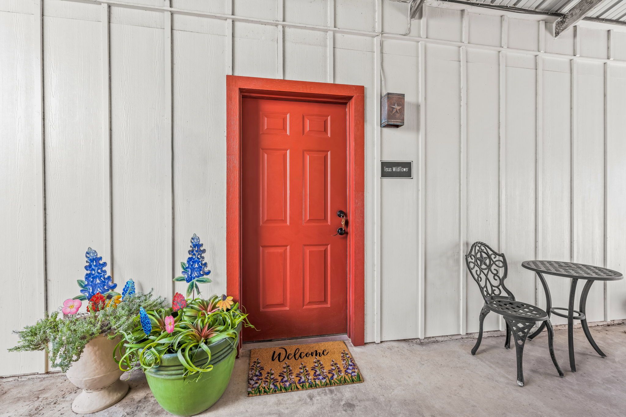 A red door with a welcome mat, a sign reading 'Texas Wildflower,' and two metal chairs and a table outside a building with white siding.