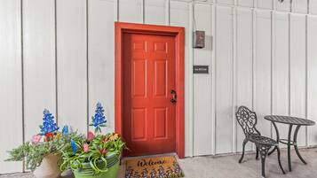A red door with a welcome mat, a sign reading 'Texas Wildflower,' and two metal chairs and a table outside a building with white siding.