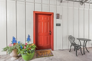 A red door with a welcome mat, a sign reading 'Texas Wildflower,' and two metal chairs and a table outside a building with white siding. - Wildflower Suite sleeps 4, by the wine trail, Luckenbach, & Fredericksburg! (Fredericksburg)