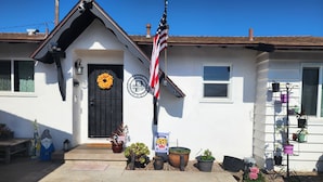 A white building with a black door, a wreath, and an American flag. There are potted plants and a metal shelf with more plants. - Charming 3-bedroom house in fabulous Oxnard with WiFi (Oxnard)