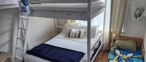 A bunk bed room with a ladder, a small bed with a Sonic the Hedgehog themed bedspread, and a window with curtains.