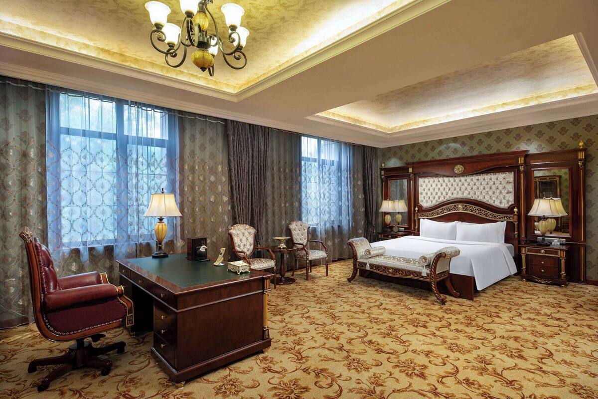 A hotel room with a large bed, a desk, two armchairs, a small table, and a nightstand with lamps.