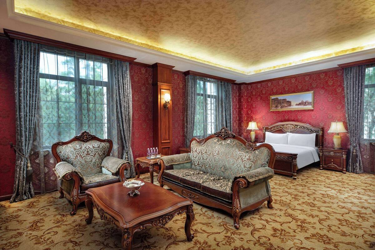 A room with a large bed, two armchairs, a wooden coffee table, and a painting on the wall.