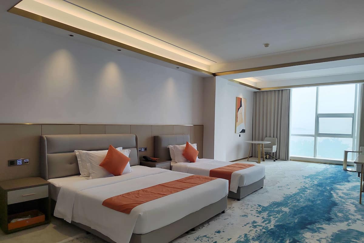 A hotel room with two beds, a small table, and a large window.