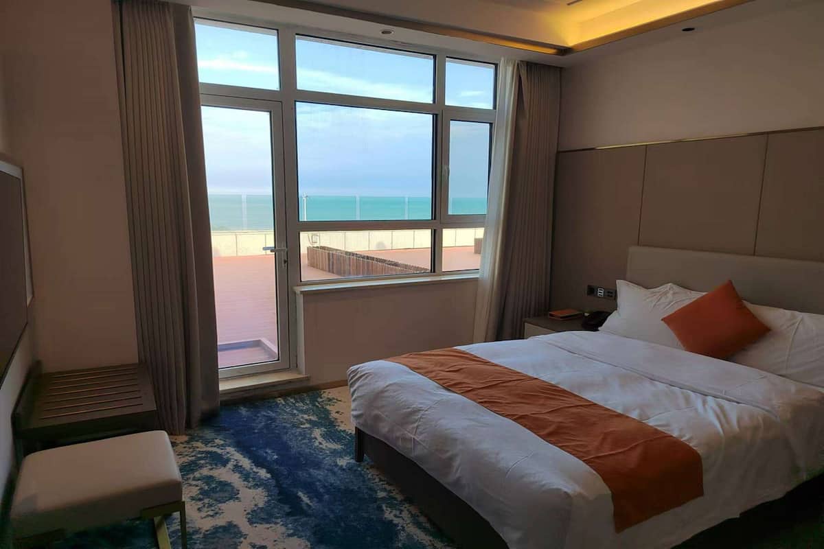A hotel room with a large window, a bed with white and orange bedding, a small bedside table, and a bench.