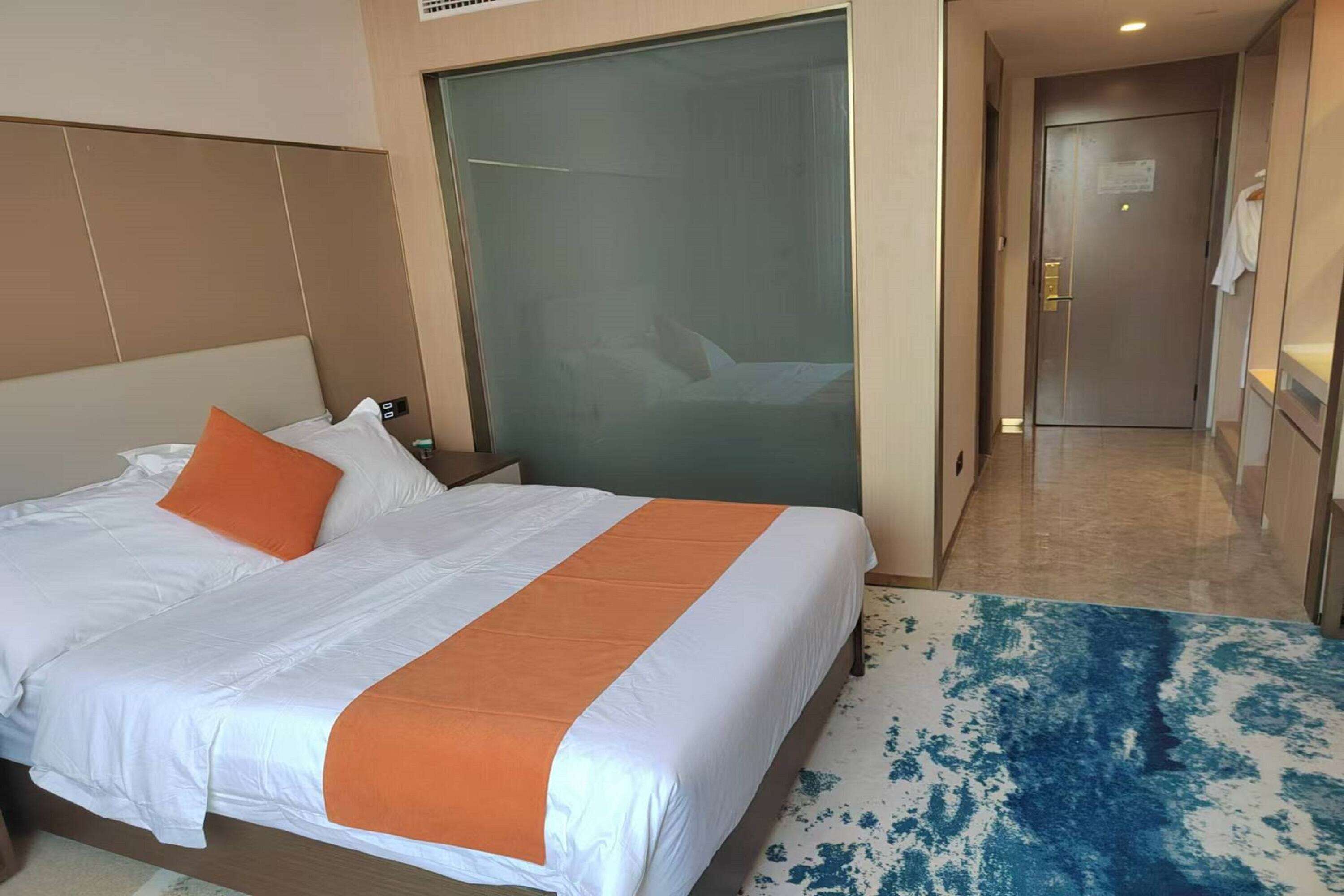 A hotel room with a large bed, a nightstand, a glass partition, and a bathroom door.