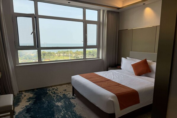 A hotel room with a large window, a bed with white and orange bedding, a nightstand, and a chair.