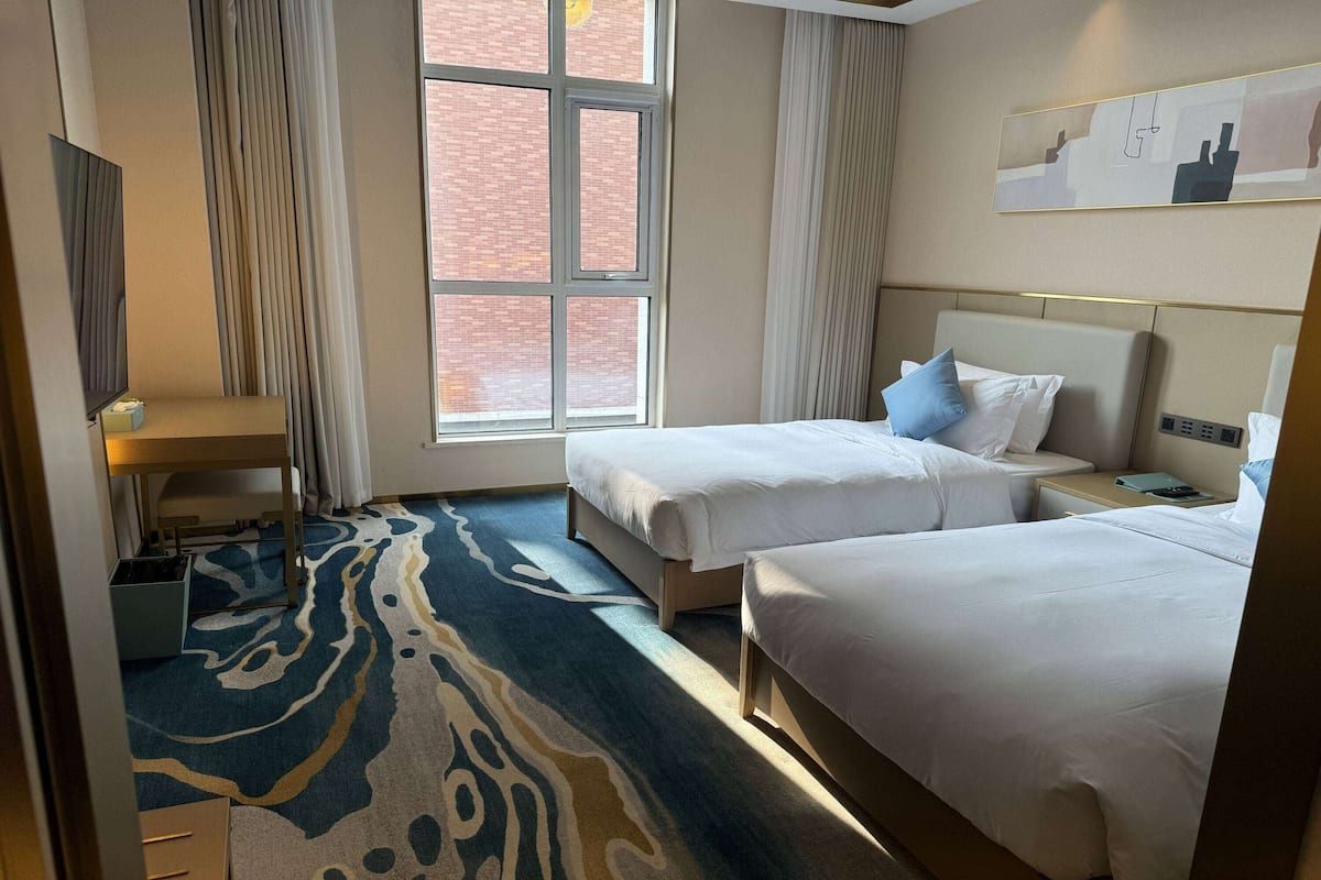 A hotel room with two beds, a large window, a patterned carpet, and a wall-mounted artwork.